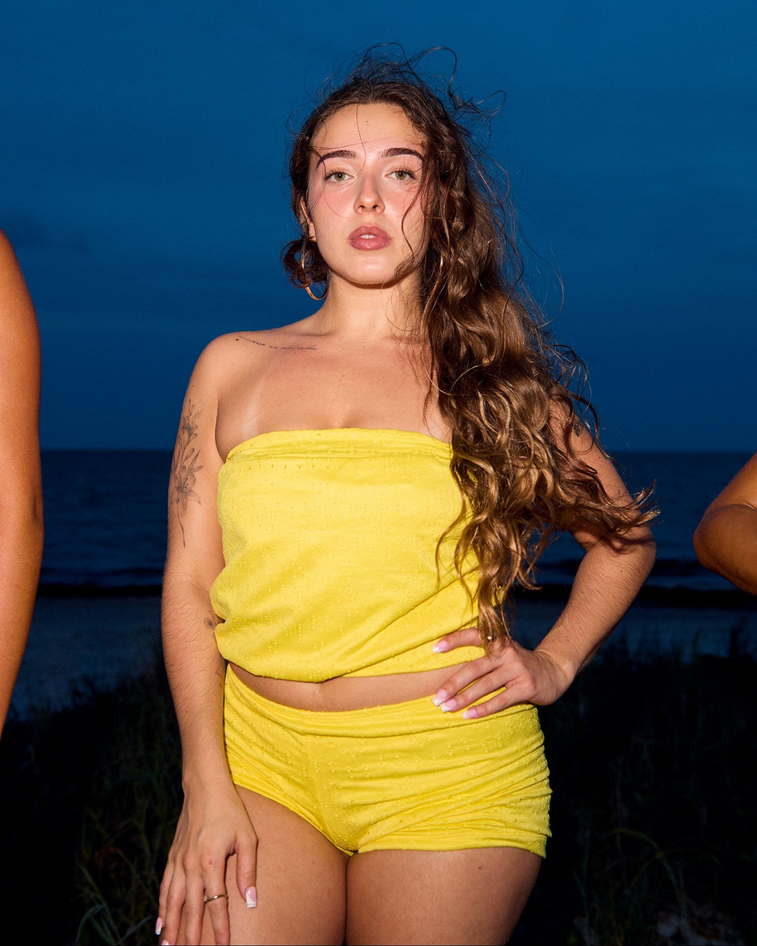 A woman standing outdoors at dusk wearing a bright yellow strapless tube top and matching shorts, with the ocean and dark sky behind her.