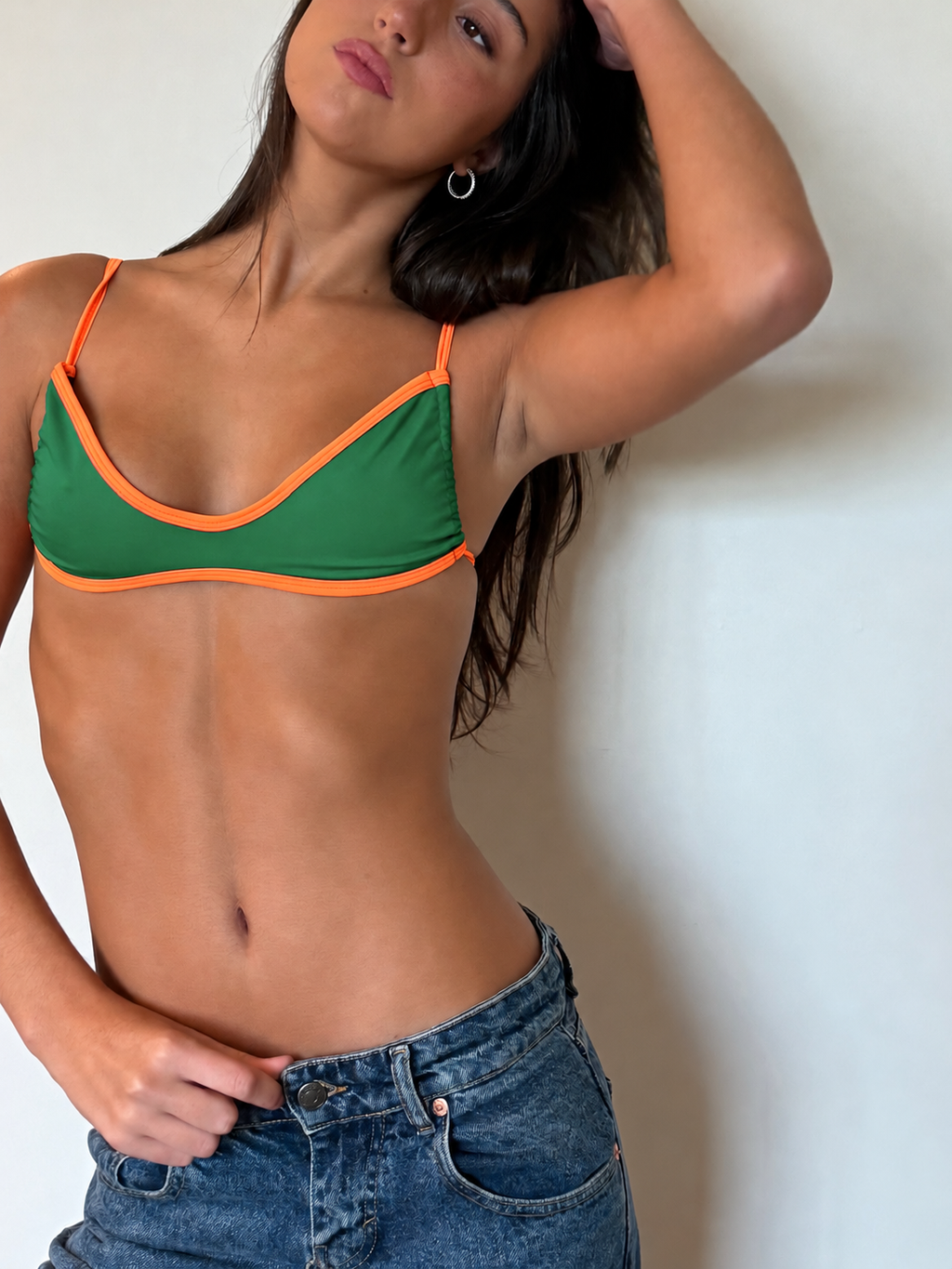 A woman wearing a green bikini top with bright orange trim, paired with blue jeans, posing against a plain white wall.